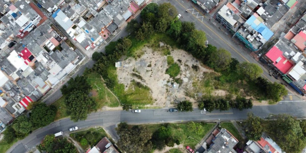 Vista aérea de un lote con árboles y suelo descubierto, rodeado por calles, viviendas y varios vehículos en circulación.