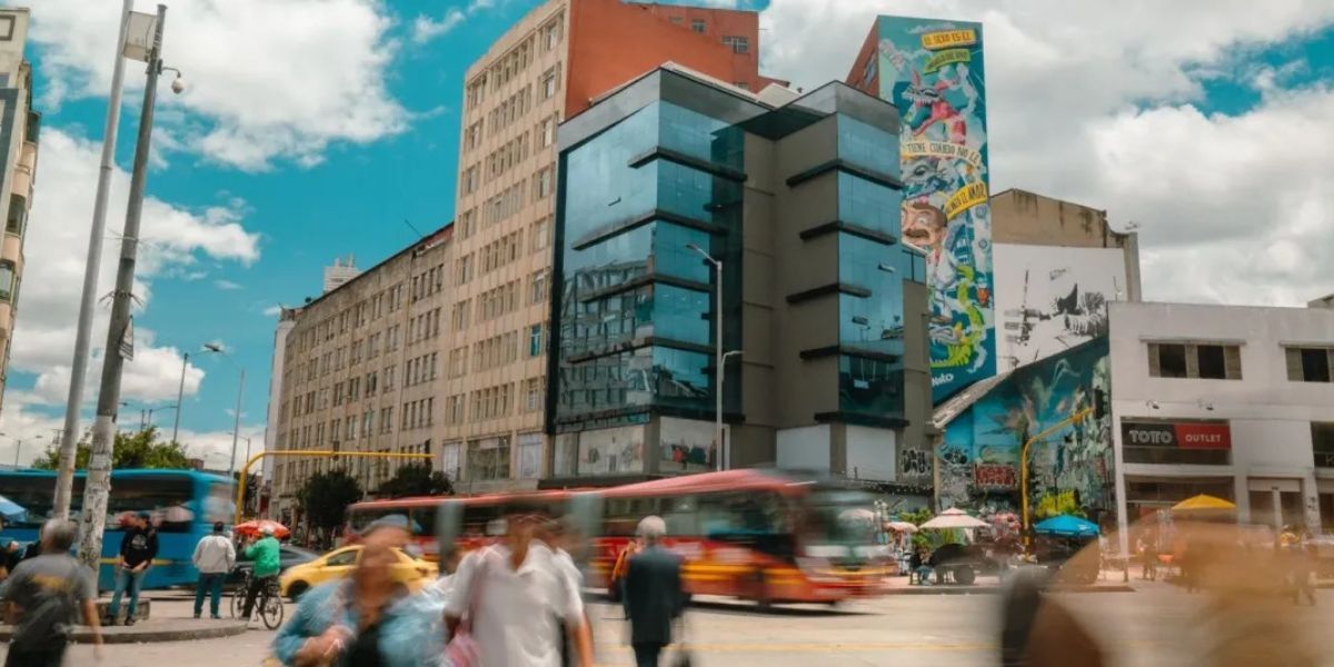 Intersección urbana con peatones y buses en movimiento. Al fondo hay edificios, uno con fachada de vidrio y murales laterales.