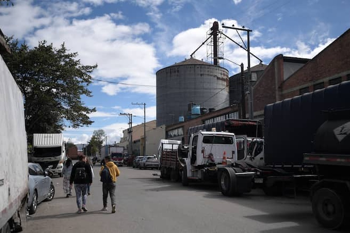 Calle industrial con varios camiones y autos estacionados a ambos lados; tres personas caminan de espaldas. Al fondo hay silos metálicos, edificios de ladrillo y postes eléctricos.		
