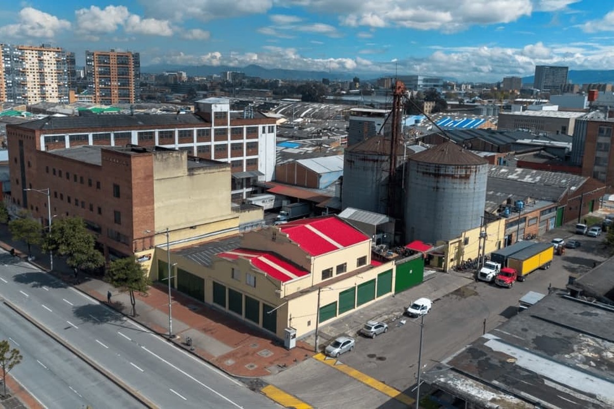 Vista aérea de zona industrial urbana: edificios de ladrillo y bodegas amarillas con techo rojo, dos grandes silos metálicos, calles amplias casi vacías y varios camiones y autos estacionados.		