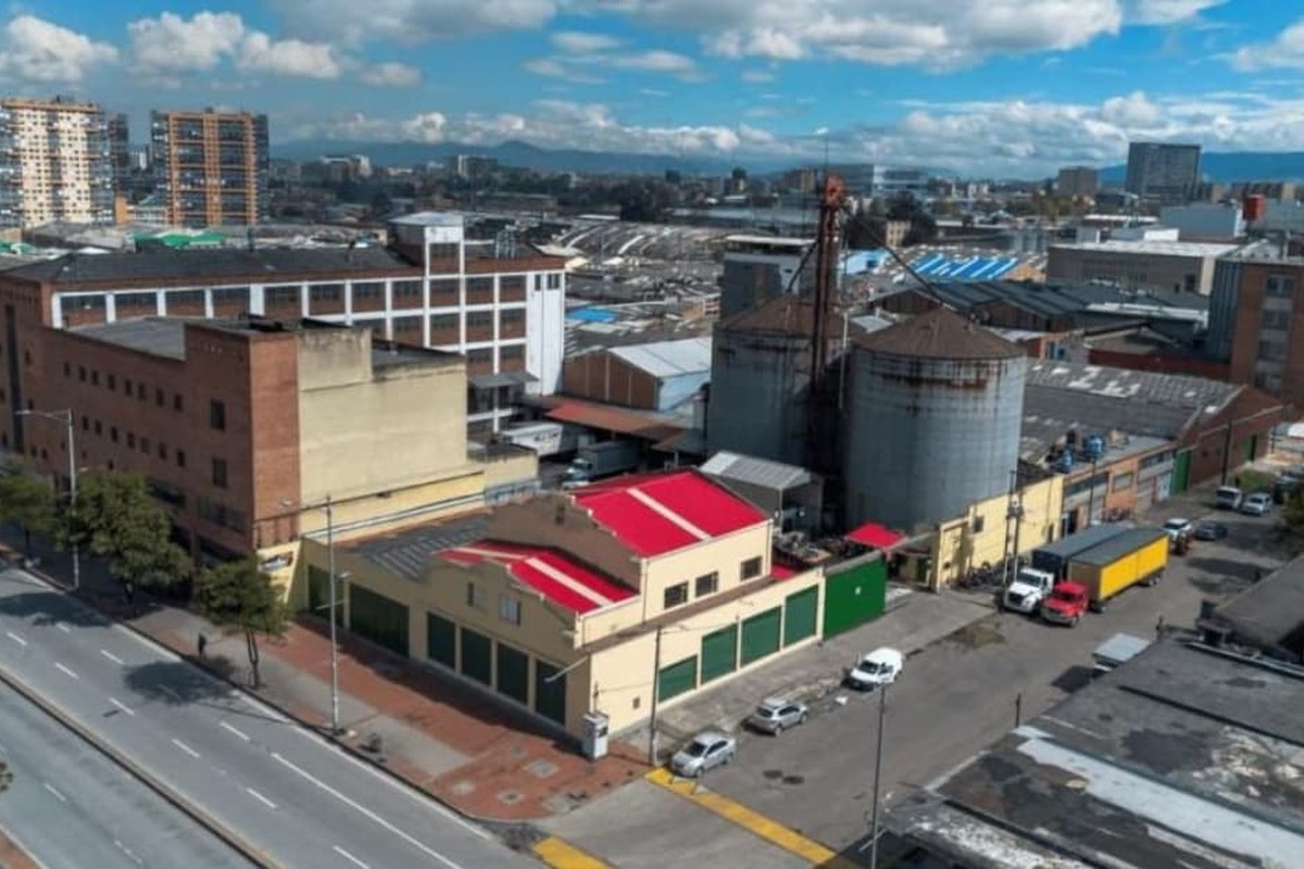 Vista aérea de zona industrial urbana con edificios de ladrillo y naves, dos grandes silos metálicos, una calle ancha casi vacía y otra con camiones y autos estacionados, cielo azul con nubes.		