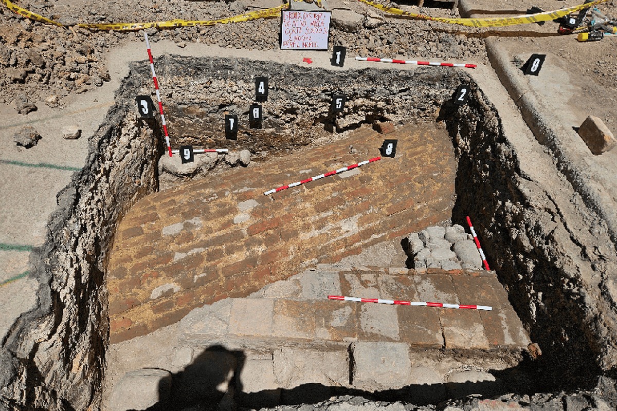 Excavación rectangular profunda con muro o bóveda de ladrillo inclinado, suelo de bloques de piedra, varas de medición rojas y blancas, etiquetas numeradas negras y cinta amarilla alrededor.		