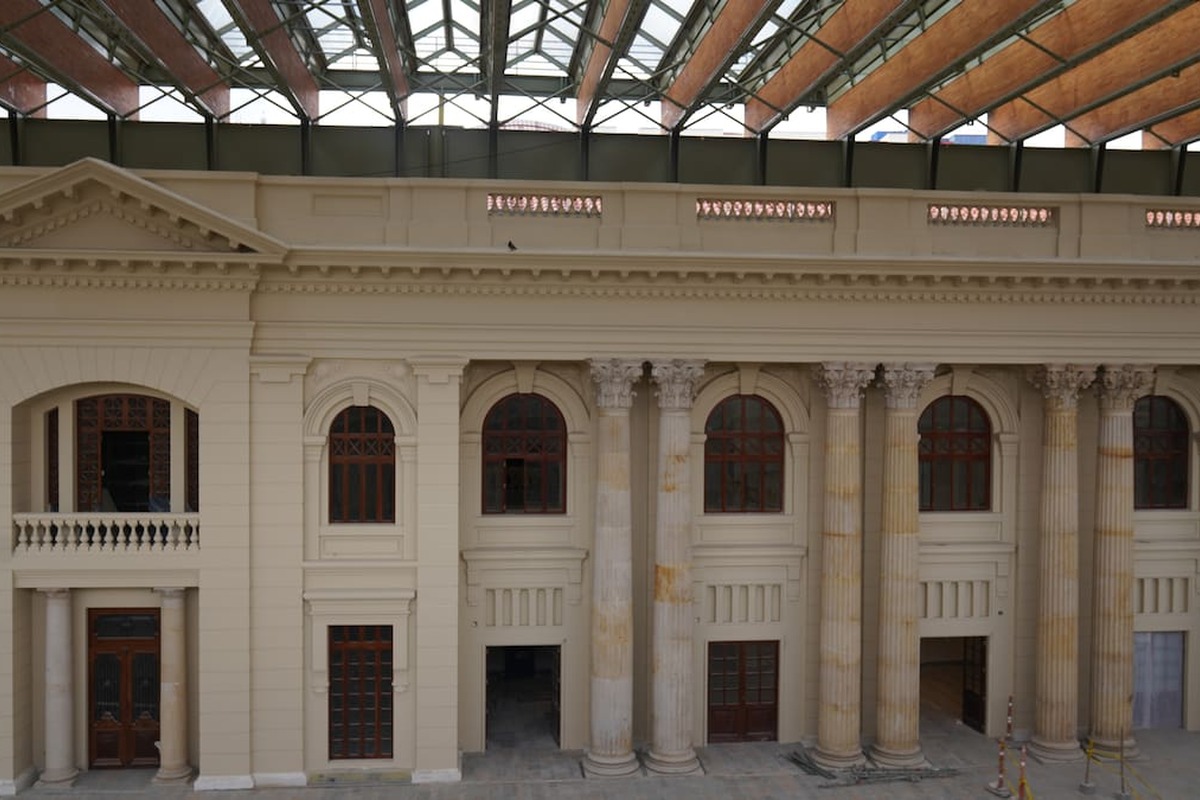 Fachada interior de edificio clásico beige con altas columnas estriadas y grandes ventanas arqueadas de madera oscura techo de vidrio y metal visible arriba sin personas ni texto.