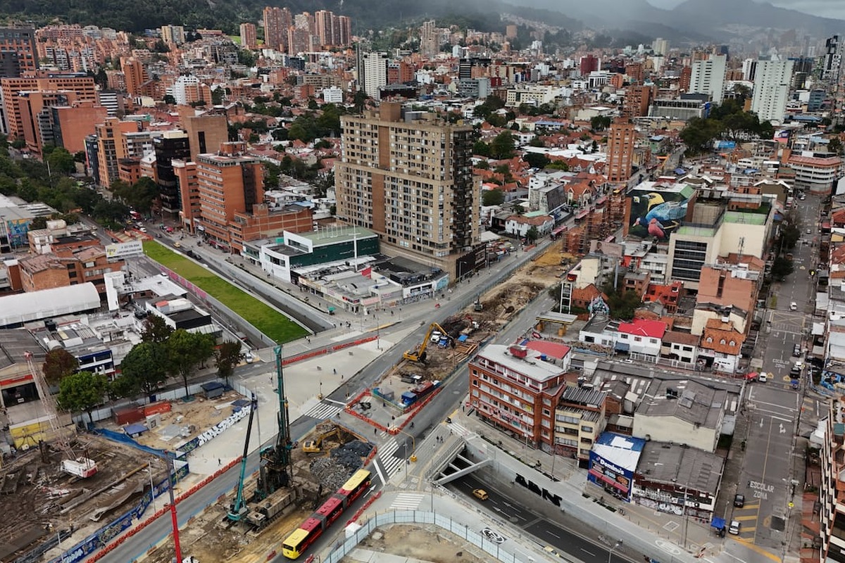 Vista aérea de ciudad con muchos edificios, calles y montañas al fondo. En primer plano, obras viales con maquinaria, buses rojos y varios peatones en aceras y zonas verdes.