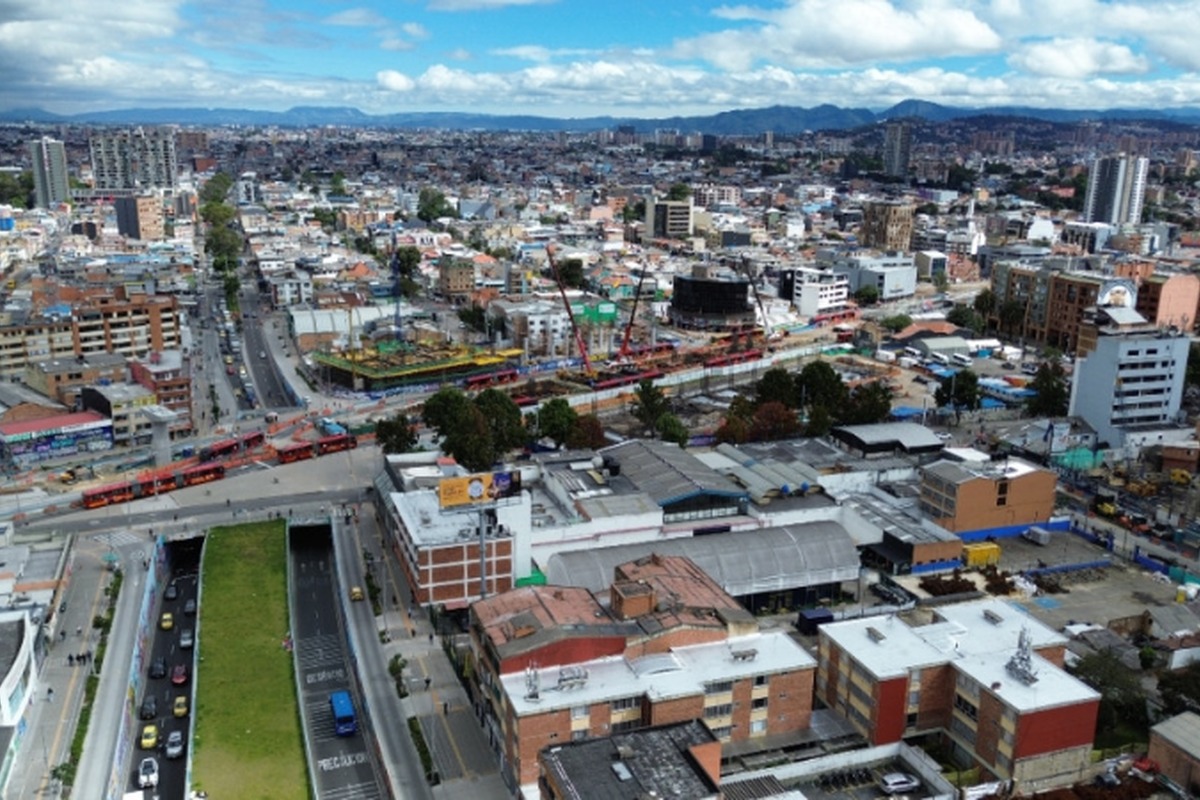 Vista aérea de ciudad densa con muchos edificios bajos y algunos altos, avenidas con autos y buses rojos articulados, obras de construcción y montañas al fondo bajo cielo nublado.