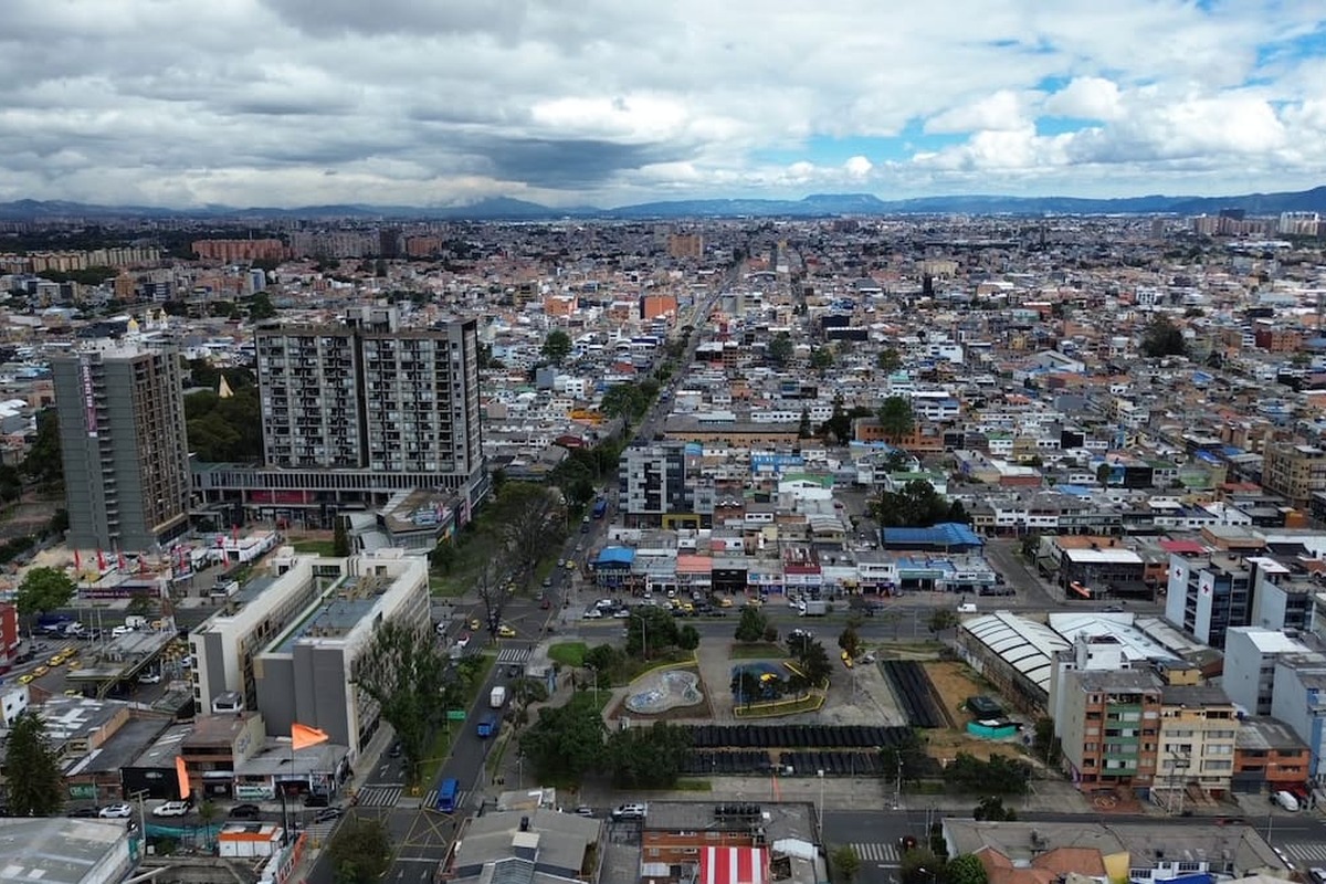 Vista aérea de una ciudad extensa con muchos edificios bajos, algunas torres altas, avenidas con autos, un pequeño parque y montañas al fondo bajo cielo nublado.