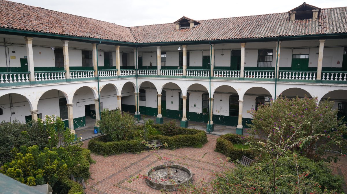 Patio interior con jardines, fuente circular, bancas y edificio de dos pisos con arcos y balcones.
