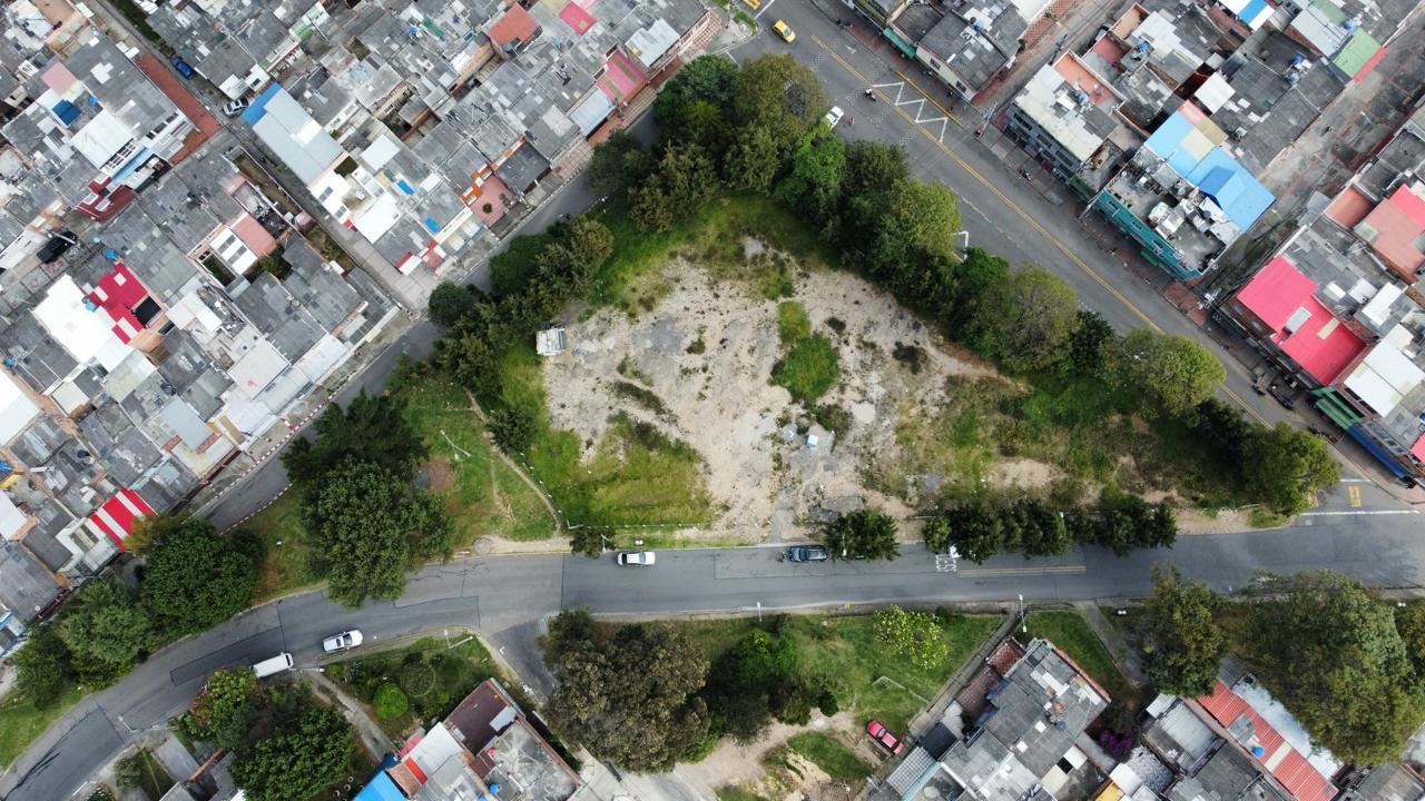 Vista aérea de un terreno baldío con árboles, rodeado de calles y viviendas; hay varios autos estacionados.