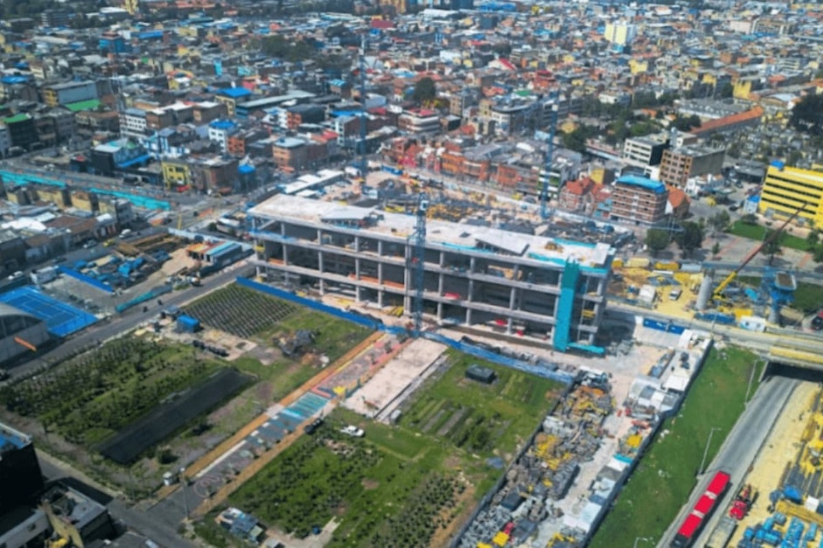 Vista aérea de ciudad densa con edificios bajos. En el centro, gran construcción gris de varios pisos en obra. Alrededor hay zonas verdes, canchas azules y vías con vehículos rojos.