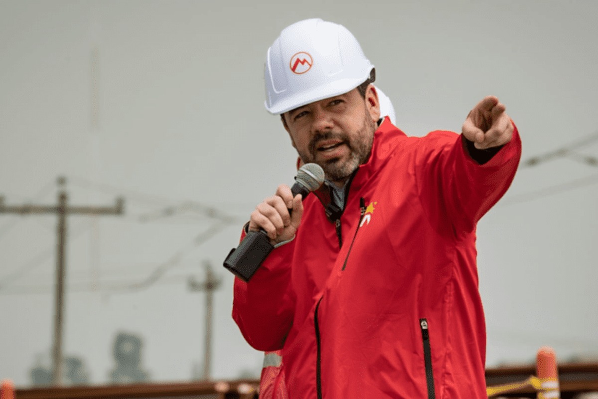 Hombre con casco blanco y chaqueta roja sostiene un micrófono y señala al frente. Fondo exterior con postes y cables eléctricos desenfocados.
