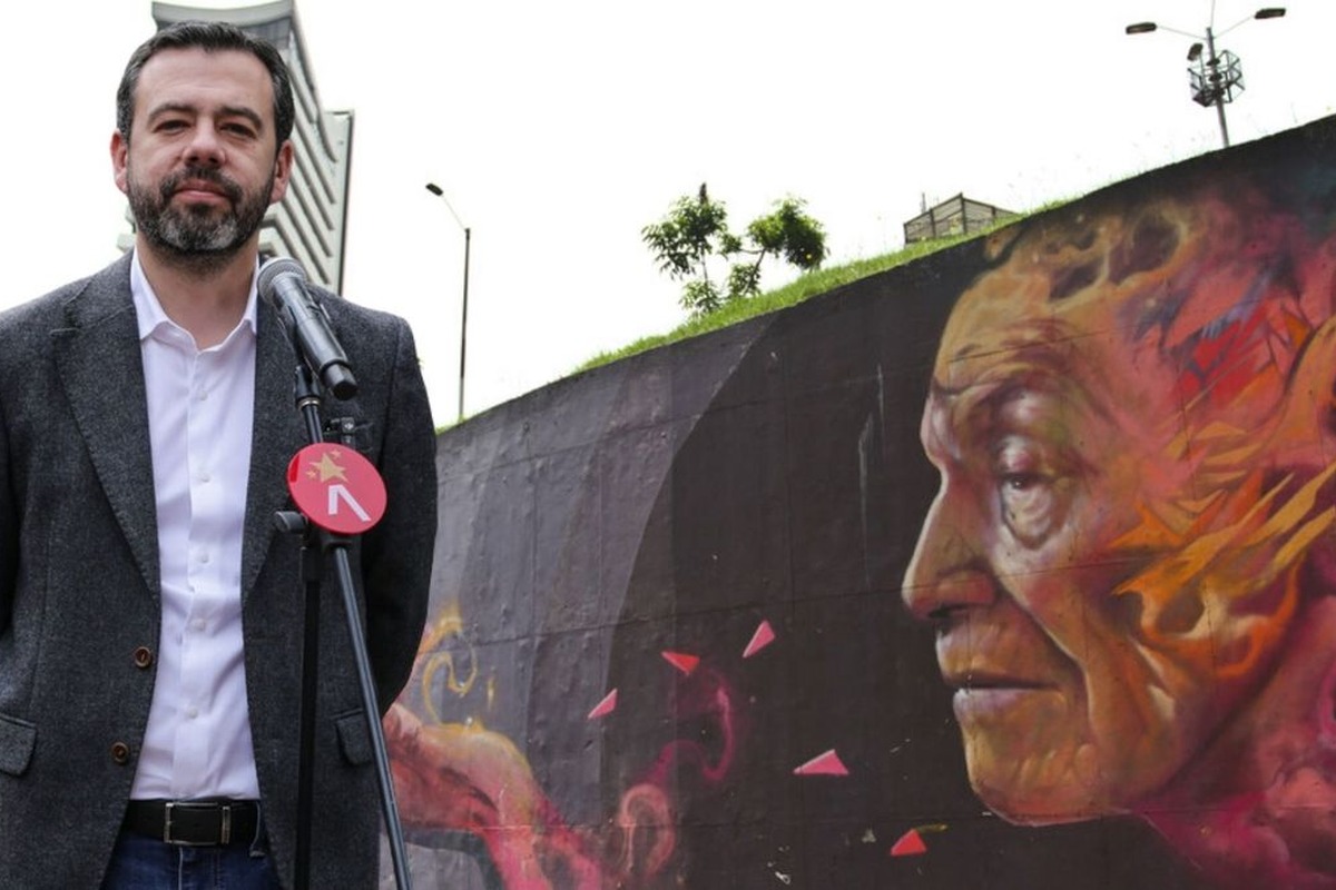 Hombre de pie ante micrófono con logo rojo y blanco, al aire libre; detrás, mural colorido del rostro de un anciano en una pared oscura junto a césped y farolas.