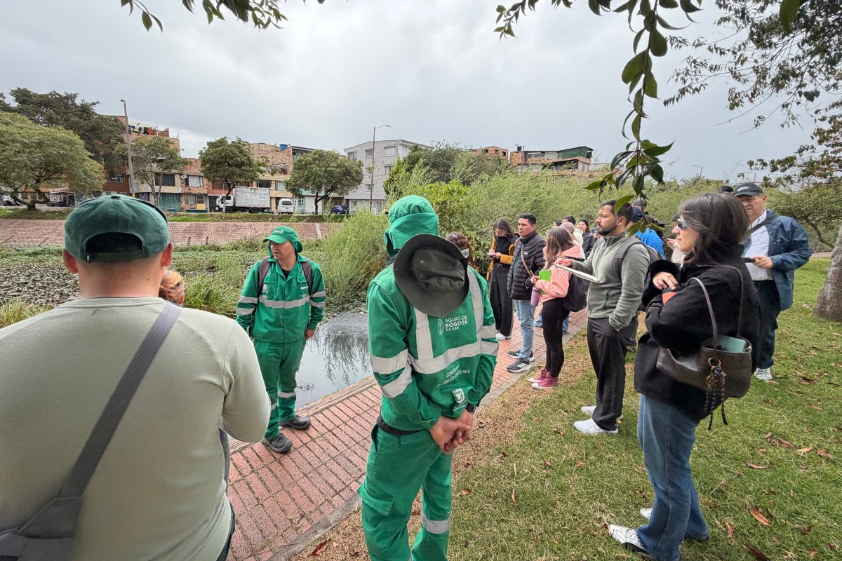 Grupo de personas junto a un canal; varias llevan uniformes verdes. Al fondo, árboles, edificios y cielo nublado