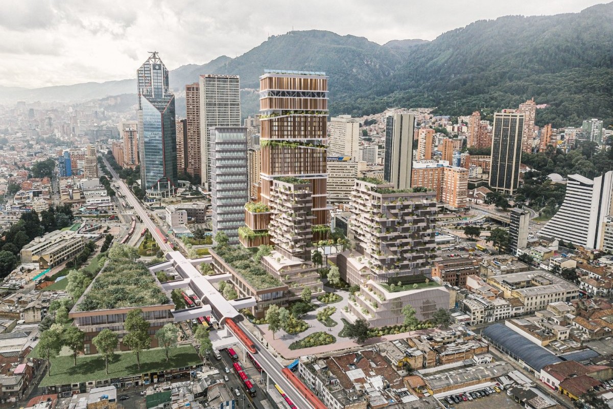 Vista aérea de edificios modernos con terrazas verdes, vías con vehículos y montañas boscosas al fondo, bajo un cielo nublado.