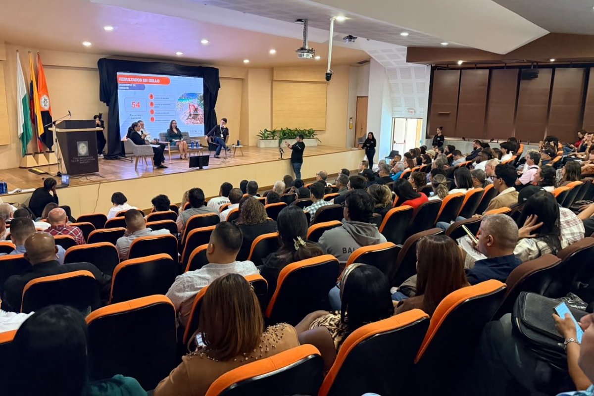 Público sentado en sillas negras y naranjas observa un conversatorio en un auditorio, con panelistas y pantalla al frente.